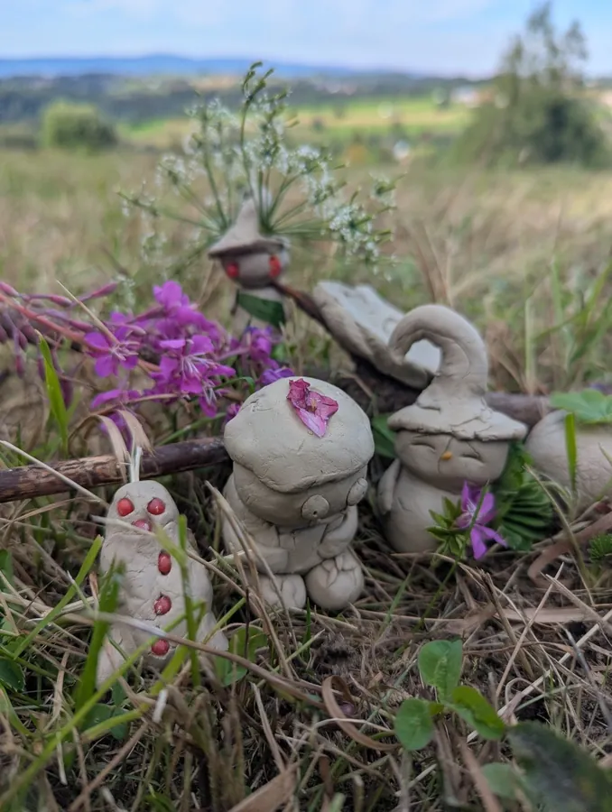 Gliniane postaci tworzące plenerową scenę z kwiatami i materiałami naturalnymi.