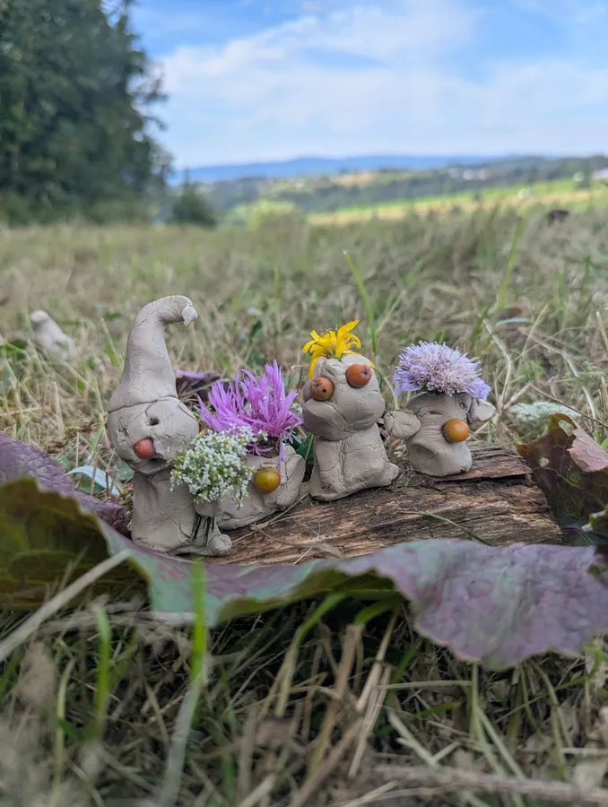 Kolorowy gliniany chór ustawiony na fragmencie drewna w otoczeniu łąki i lasu.